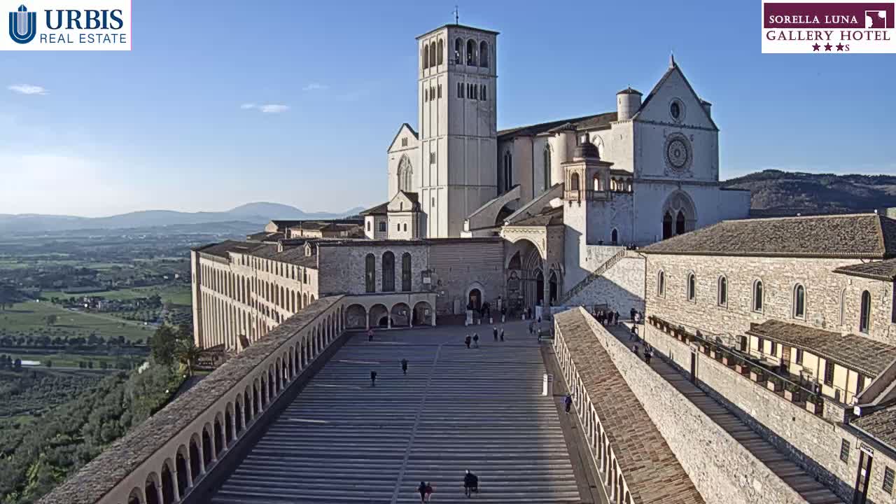 A large stone basilica and adjoining buildings are situated on a hill overlooking a valley under a clear, sunny sky.