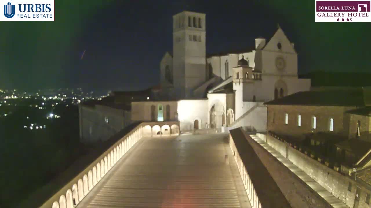A nighttime view of a large, illuminated stone building complex, possibly a church or monastery, overlooking a city at night.