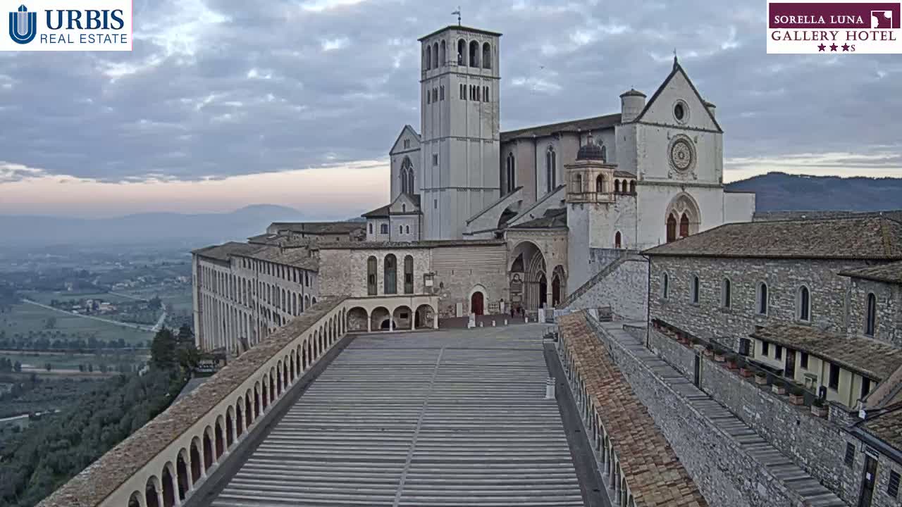 A large stone basilica overlooks a long, paved plaza and surrounding buildings, under a cloudy but bright sky.