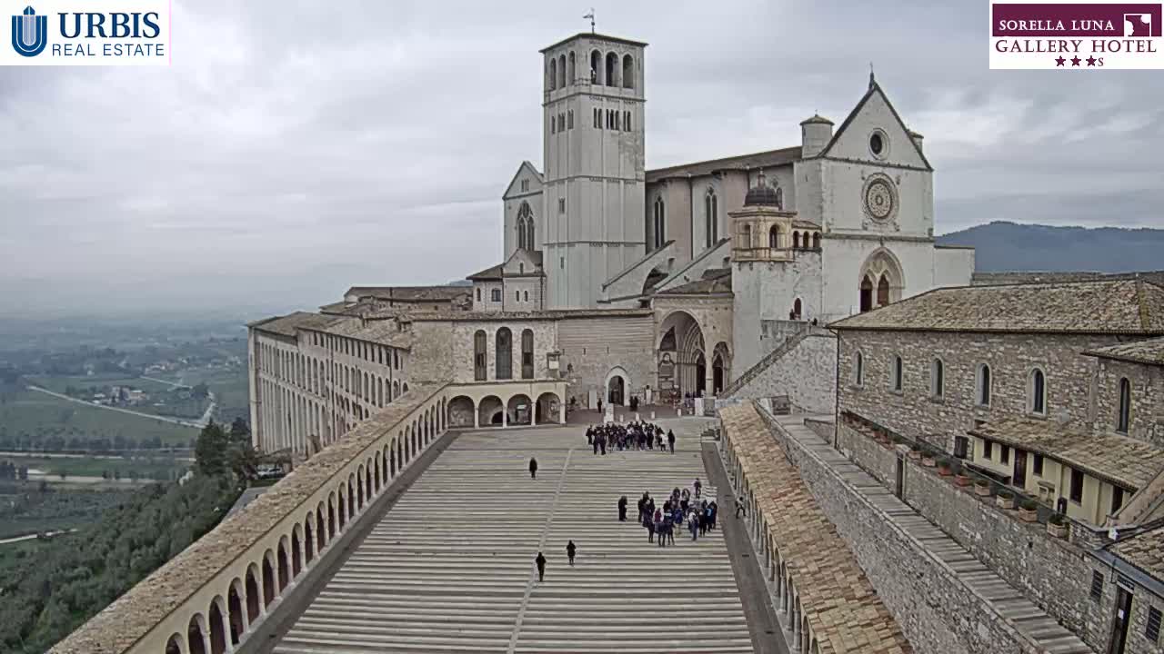 An aerial view shows a large, light-colored basilica and adjoining buildings on a hilltop under an overcast sky, with a wide paved plaza between them and several people walking about.