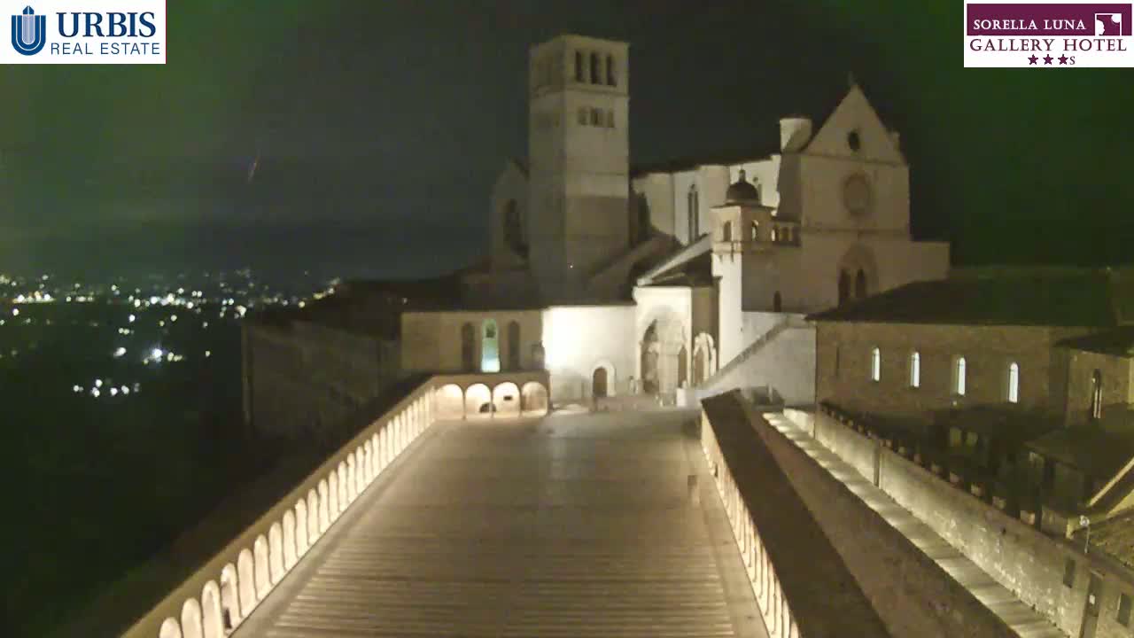 A large, illuminated stone structure at night, overlooking a city with distant lights under a dark, possibly cloudy sky.