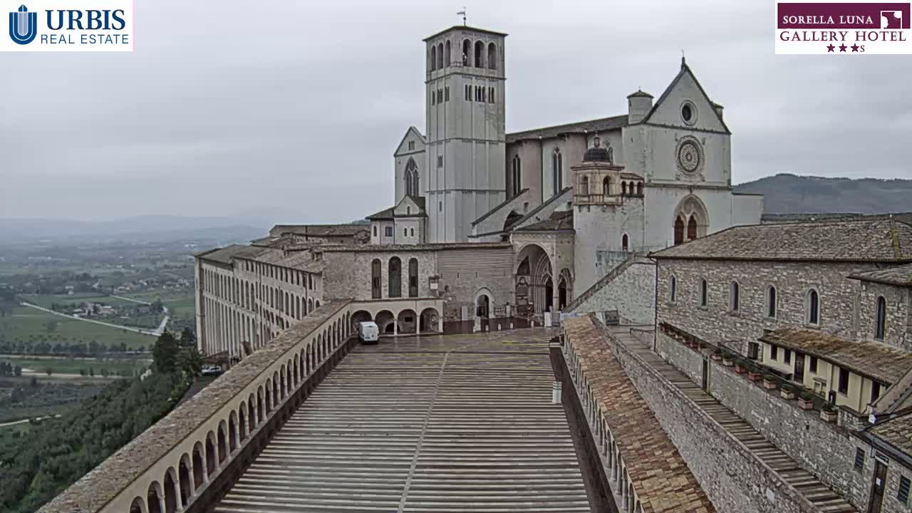 A large stone basilica and adjoining buildings overlook a valley on an overcast day.