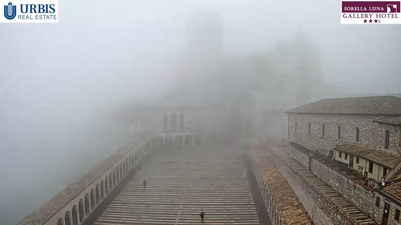 A long, stone staircase and arched walkways are shrouded in dense fog, with stone buildings visible on either side.