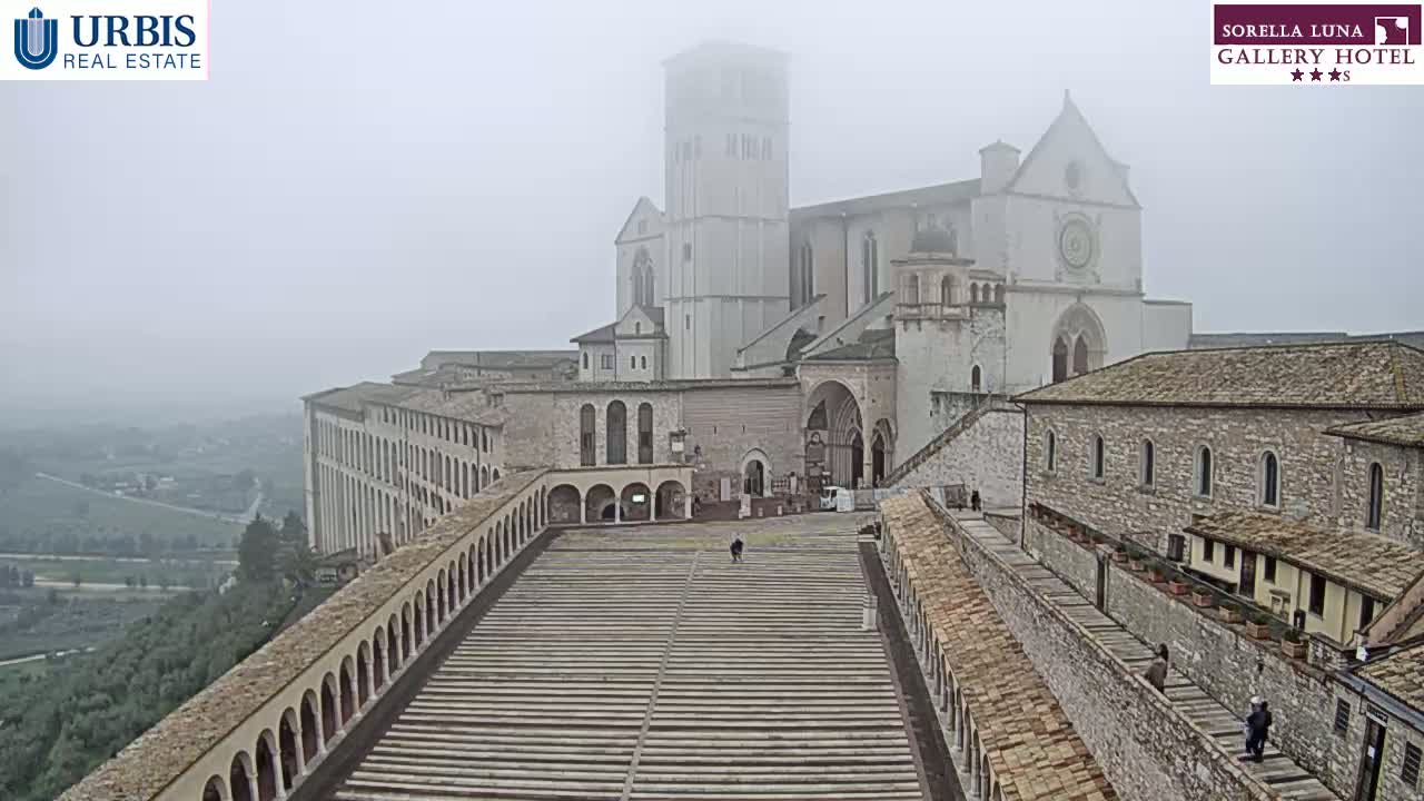 A large stone basilica and adjacent buildings are visible through a thick fog, overlooking a long, sloped plaza with arched walkways on either side.
