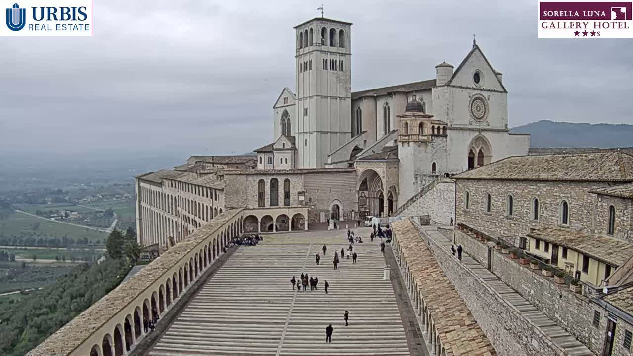 A large stone basilica and surrounding buildings are situated on a hill overlooking a valley under an overcast sky.