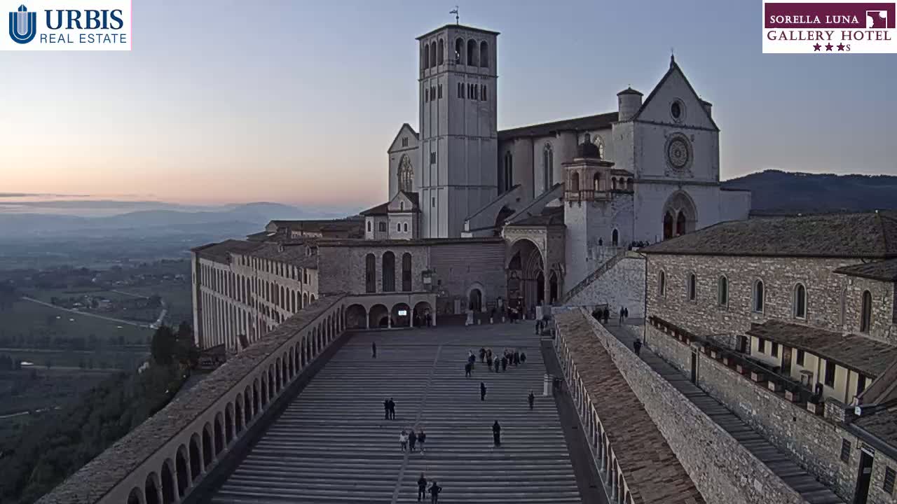 An expansive stone plaza with a large basilica and surrounding buildings is viewed from above during a dusky, twilight sky.