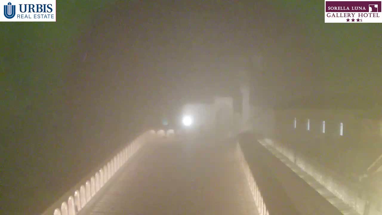 A long, dimly lit pathway with low walls on either side is shrouded in thick fog at night.