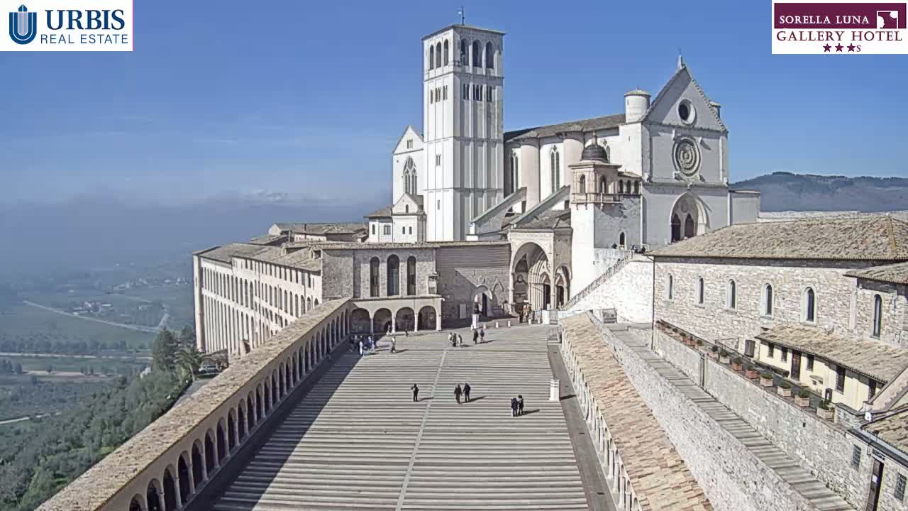 A large stone basilica and adjoining buildings sit atop a hill overlooking a valley on a clear, sunny day.