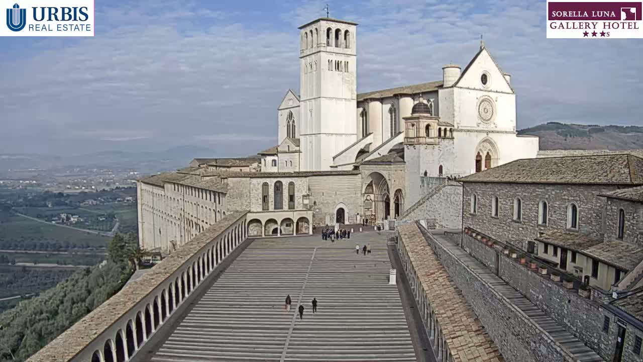 A large, light-colored basilica stands on a hilltop overlooking a valley under a mostly clear sky, with a long, wide staircase leading to its entrance.
