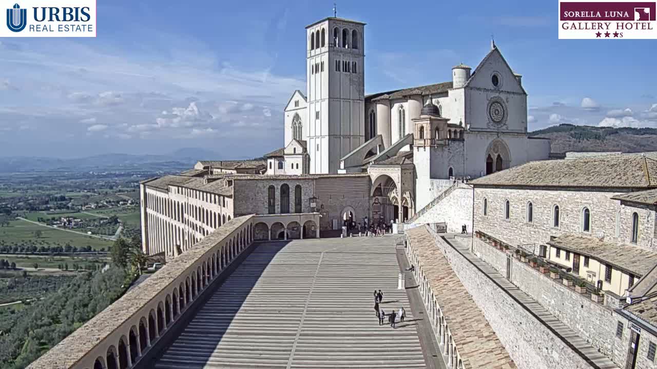 A large, light-colored stone basilica complex overlooks a sunny valley, with a wide plaza leading to the main entrance.