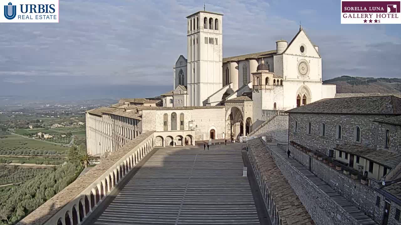 A large, light-colored basilica with a tall bell tower overlooks a wide, descending staircase and a sprawling landscape under a mostly clear, sunny sky.
