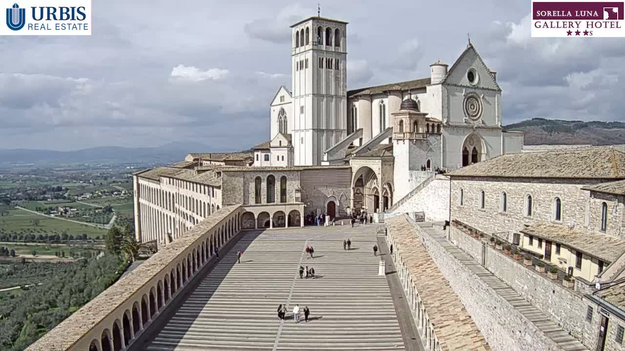 A large, light-colored basilica and adjoining buildings are seen from above on a partly cloudy day, with a wide plaza in the foreground and a hilly landscape visible in the distance.
