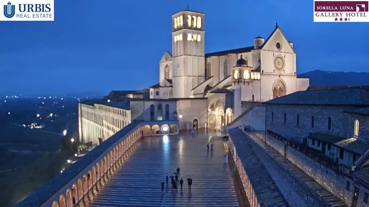 A large, illuminated basilica is situated at the top of a long, descending staircase at twilight, with a few people walking on the stairs.