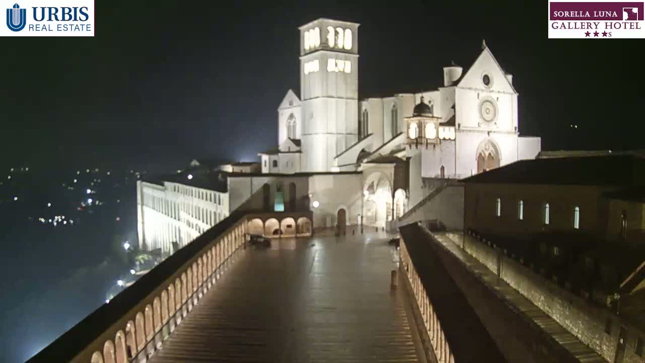 A large, illuminated basilica stands at night, viewed from an elevated position, with a wet, paved area in the foreground.