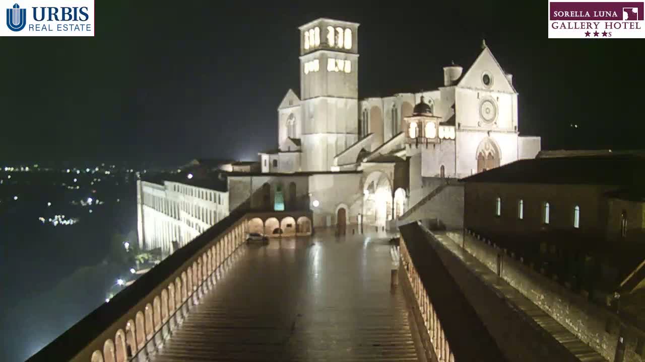 A large, illuminated stone basilica sits atop a hill at night, overlooking a city with wet, paved pathways leading down to it.