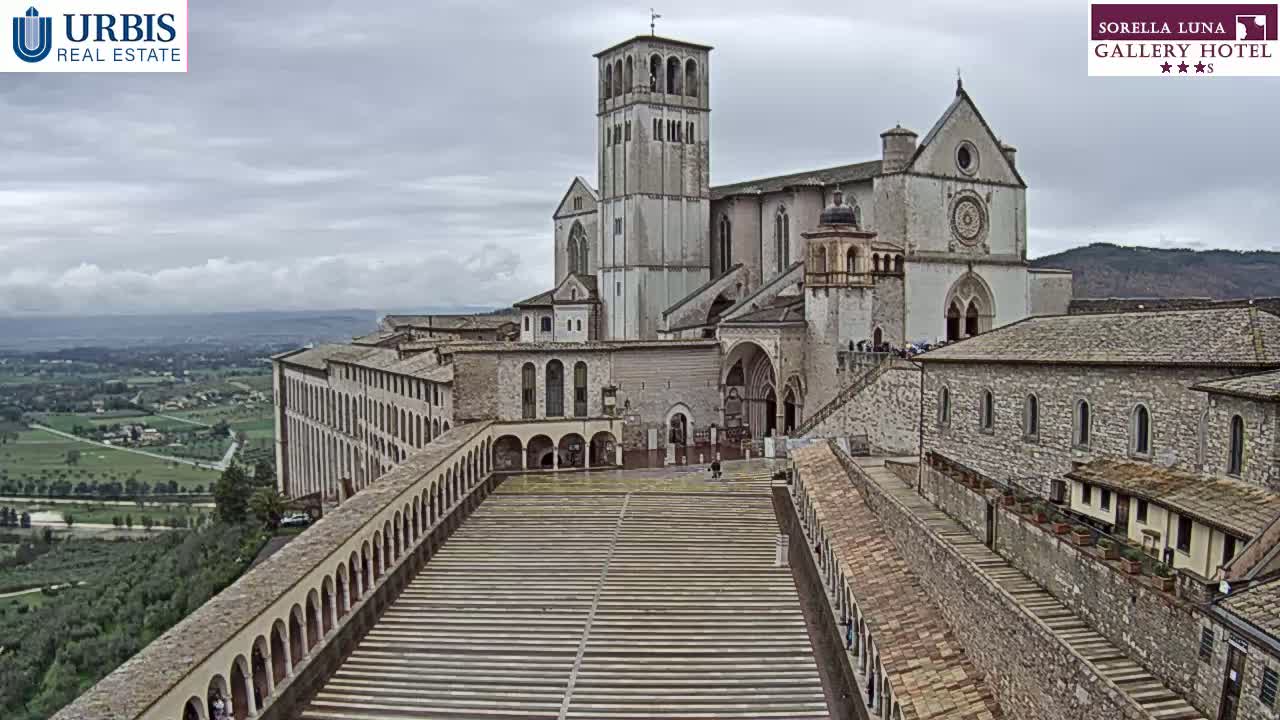 A large stone basilica overlooks a long, tiered plaza, with a sprawling landscape visible in the background under an overcast sky.