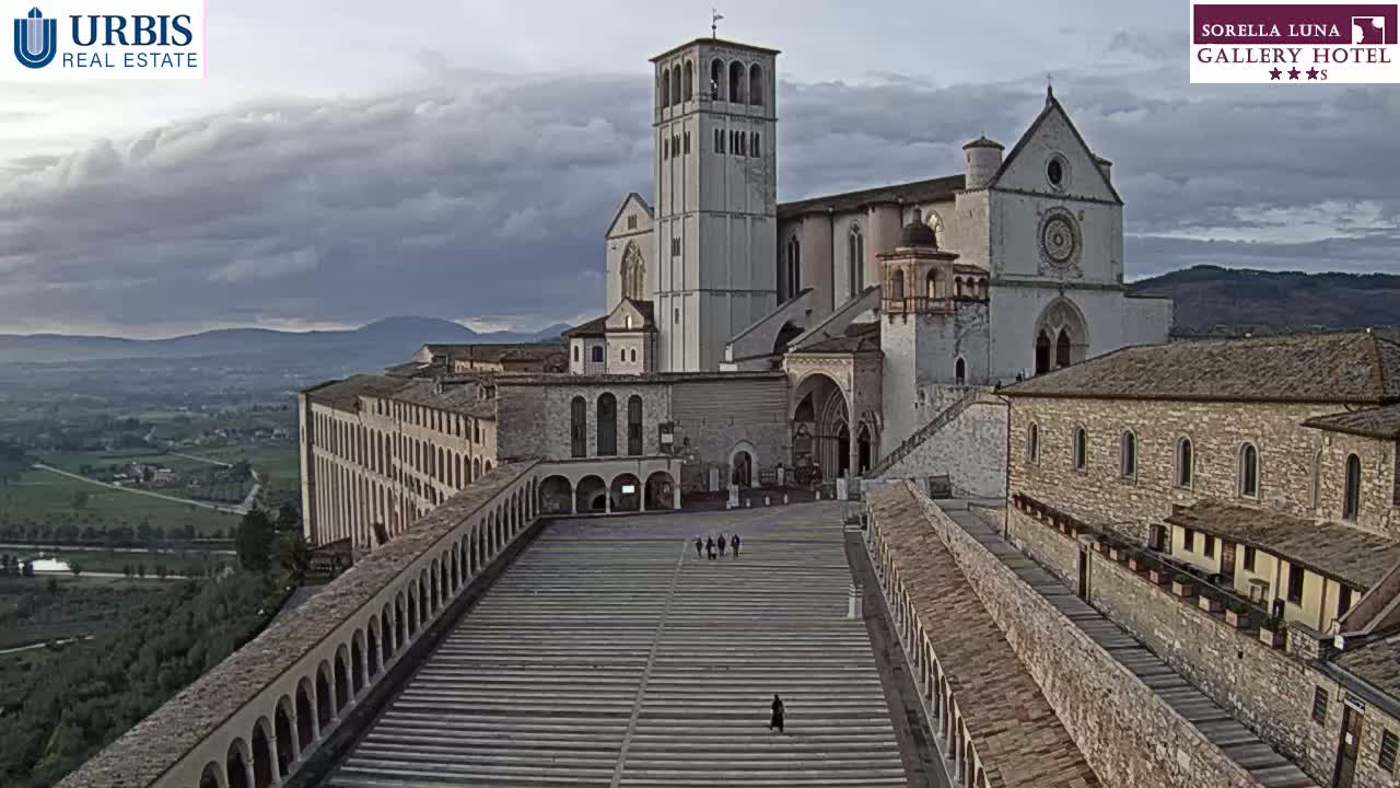 A large, light-colored basilica complex stands on a hill overlooking a verdant valley under a cloudy sky.