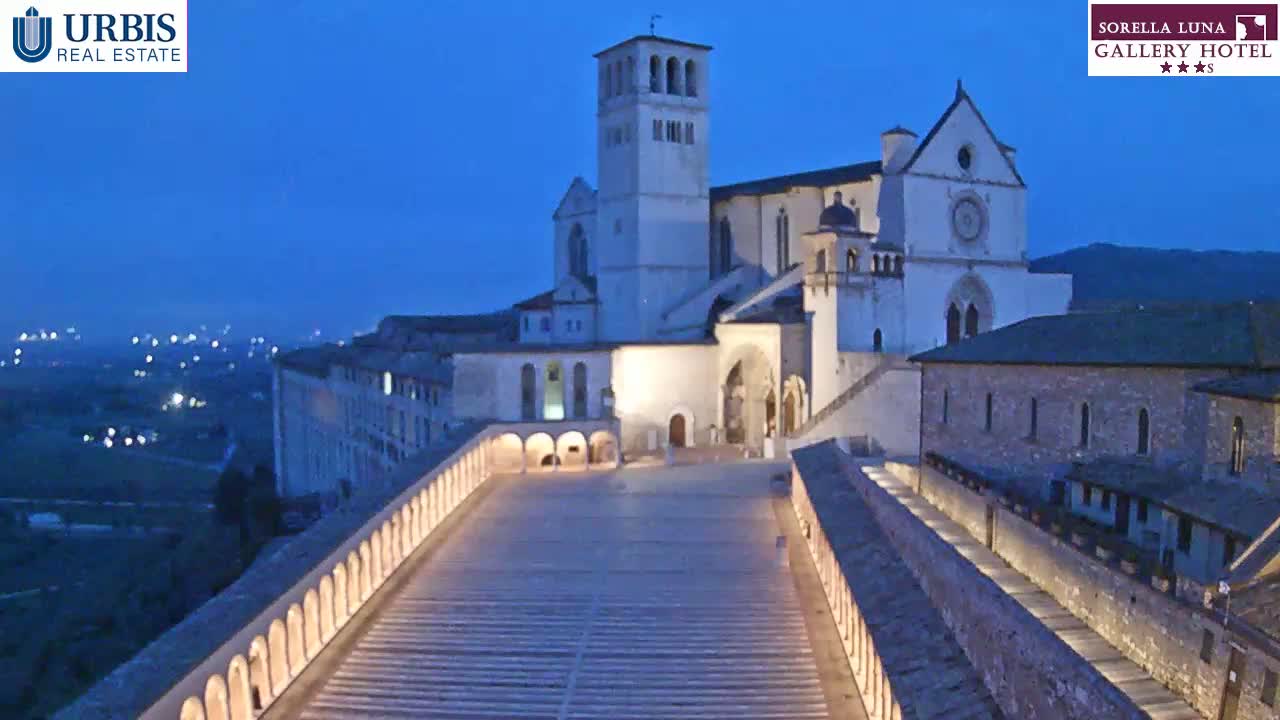 A large, illuminated basilica sits atop a hill at dusk, overlooking a city with a long, arched walkway extending from the building.