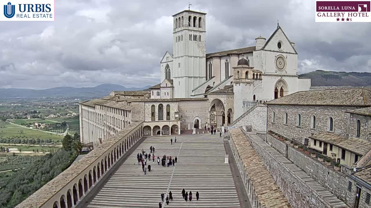A large, light-colored basilica complex with surrounding buildings and a wide, stepped plaza is seen on an overcast day, overlooking a verdant valley.