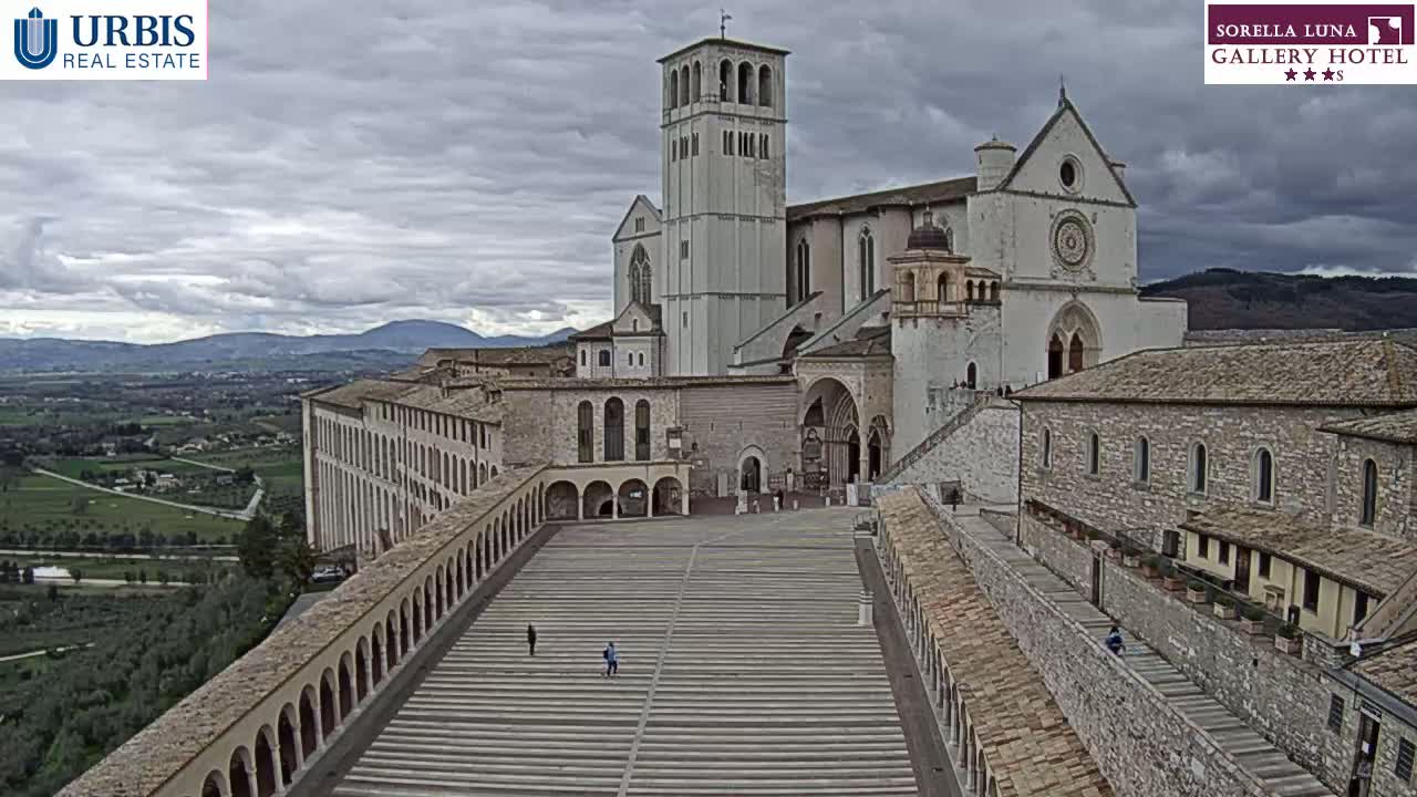 A large stone basilica and adjoining buildings are situated on a hill overlooking a valley under an overcast sky.