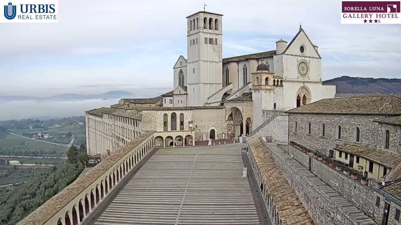 A large, light-colored stone basilica with a tall bell tower overlooks a wide, paved plaza flanked by arched walkways, all under a mostly cloudy sky with a low-lying fog in the distance.