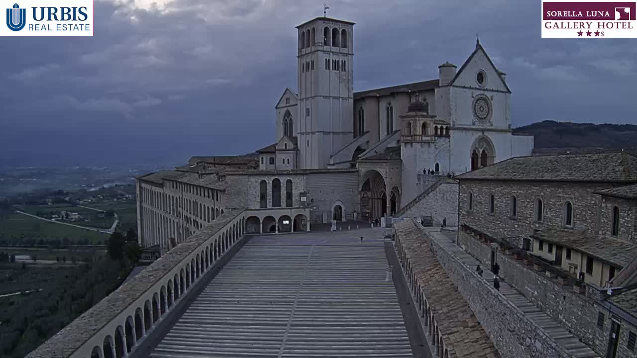 A large stone basilica with a tall bell tower is situated on a hill overlooking a valley under an overcast sky.