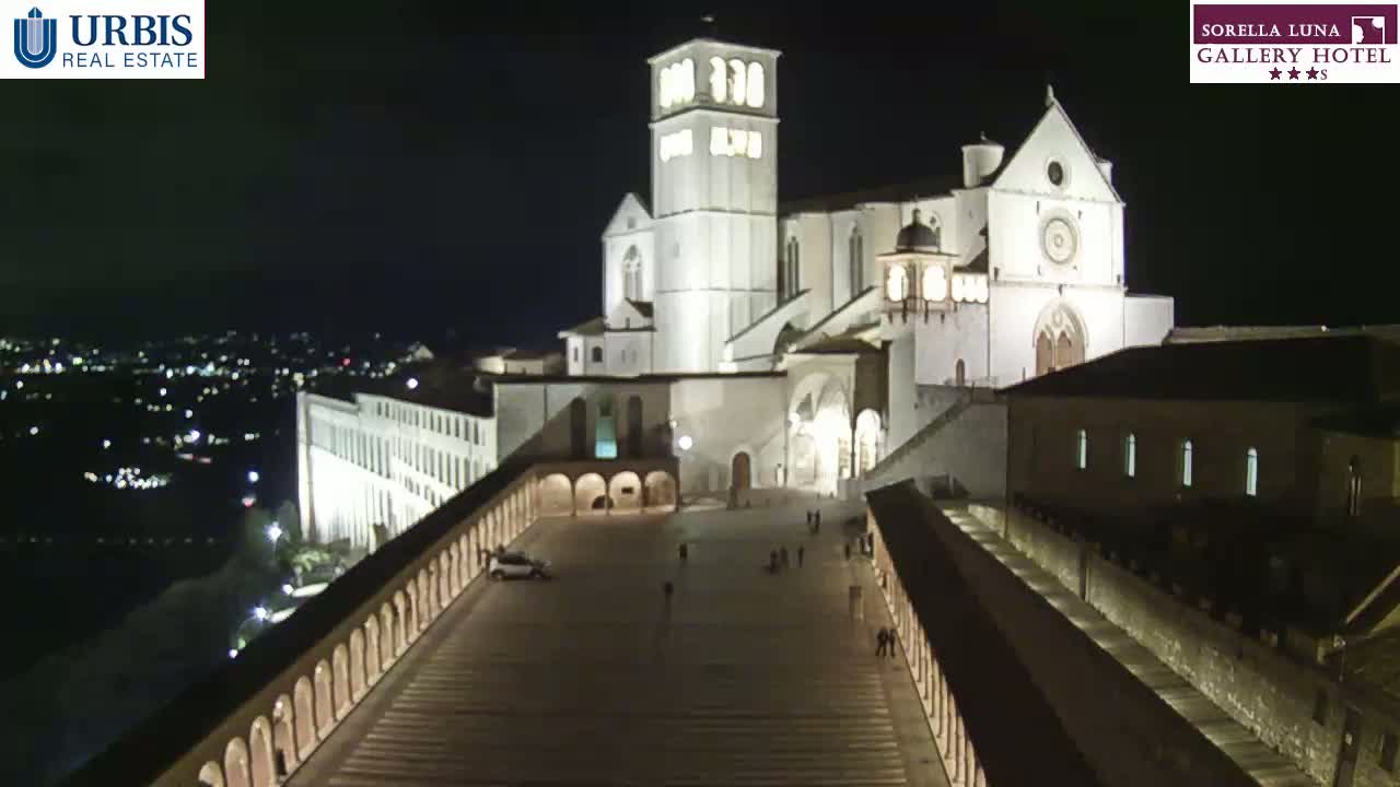 At night, a large, illuminated basilica stands prominently, surrounded by a plaza and other buildings, overlooking a city speckled with lights.