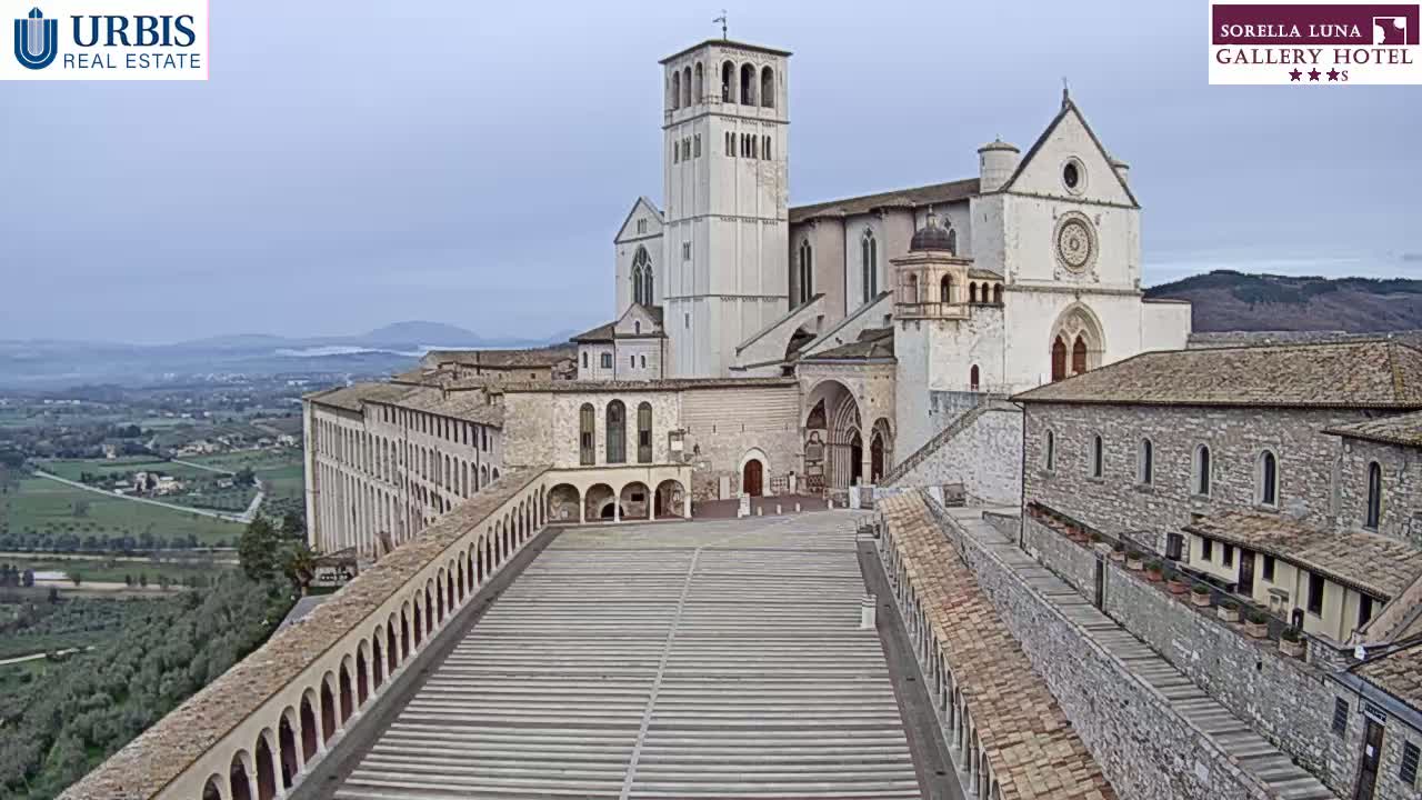A large stone basilica and adjoining buildings overlook a paved plaza, all under a cloudy sky.