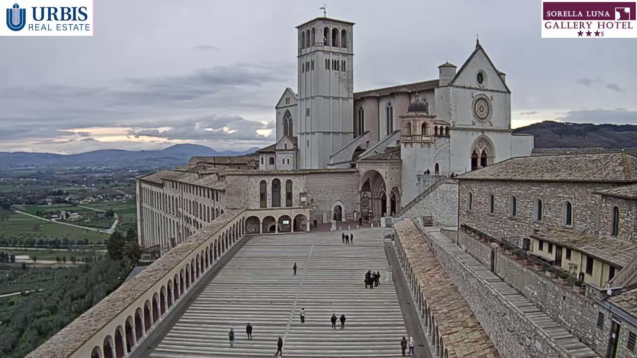 A large stone basilica and adjoining buildings are viewed from above on an overcast day, with a wide plaza and long staircase leading to the main entrance.