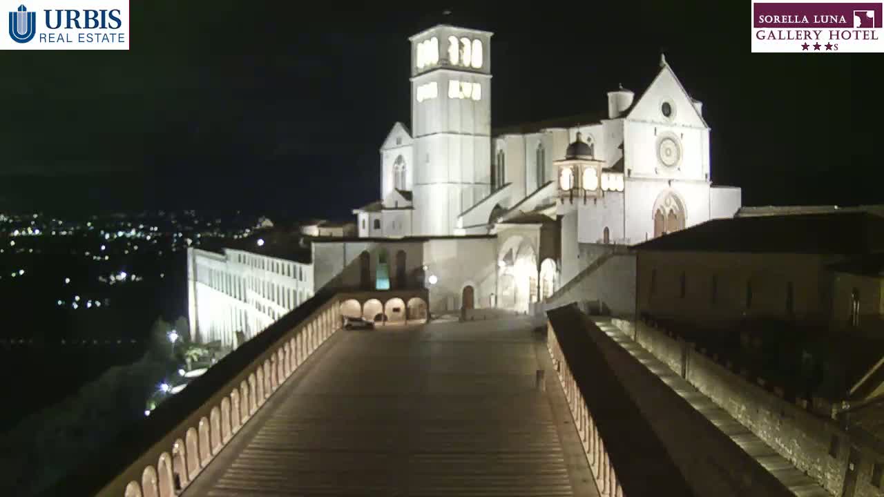 A large, illuminated basilica stands on a hilltop at night, overlooking a city with a long, arched walkway leading down to it.
