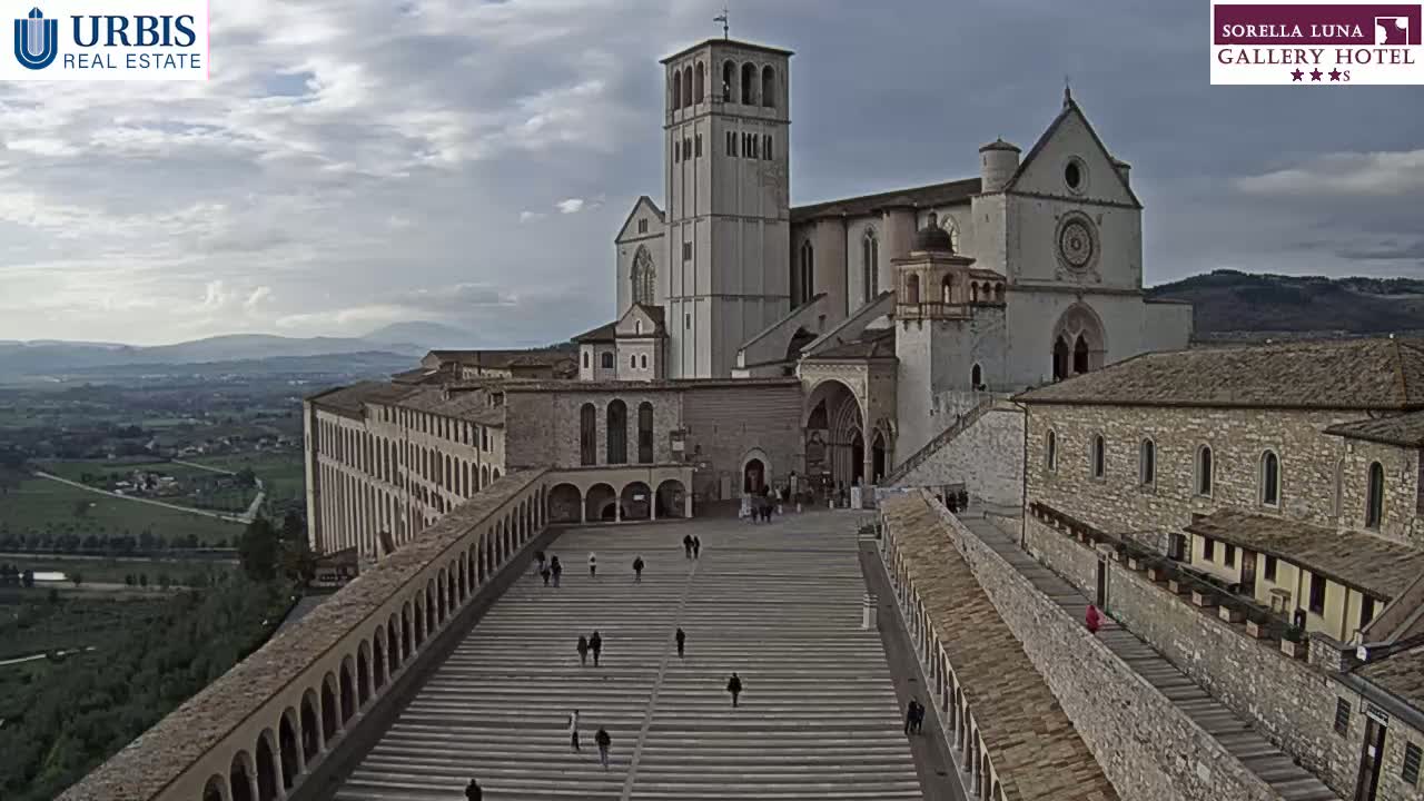 A large stone basilica and surrounding buildings are seen from above on a partly cloudy day, with a wide staircase leading to the main entrance.