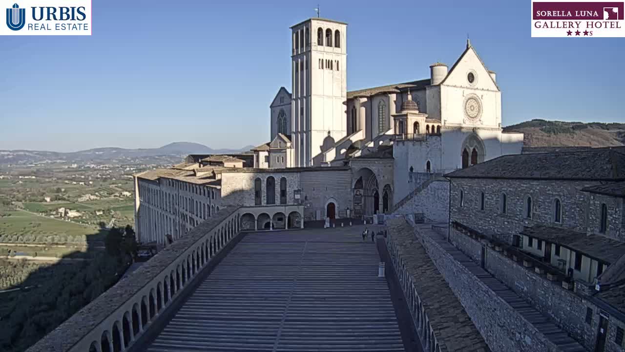 A large stone basilica and surrounding buildings are viewed from above on a clear, sunny day, overlooking a valley in the distance.