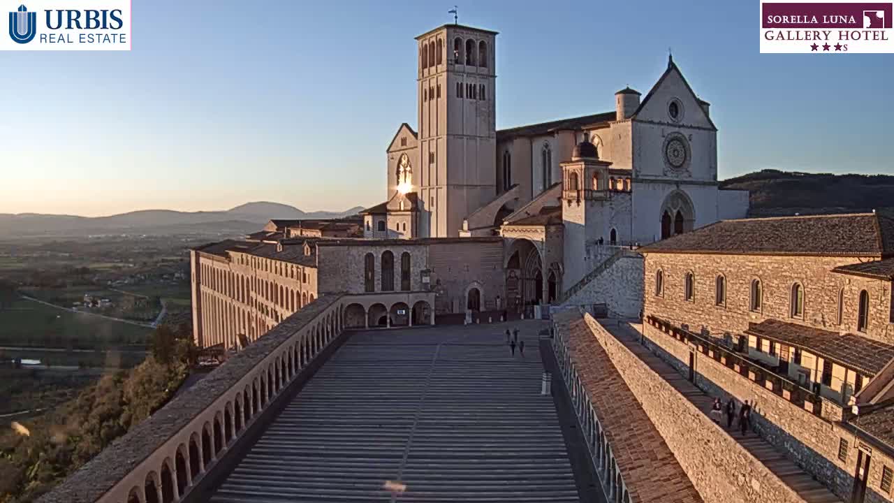 A large stone basilica and its surrounding buildings are shown at sunset, overlooking a valley.