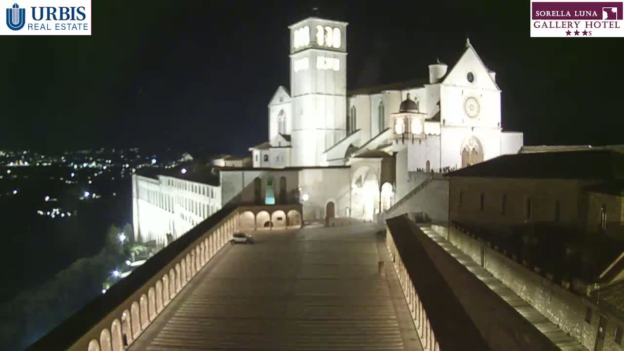 A large, illuminated basilica stands at night on a hill overlooking a city, with a long, paved walkway leading down to it.
