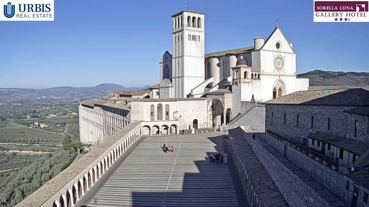 A large, light-colored basilica with a tall bell tower stands on a hill overlooking a valley under a clear blue sky.