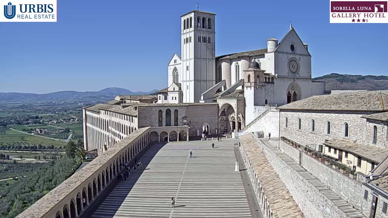 A large, light-colored basilica and adjoining buildings are situated on a hill overlooking a valley on a sunny day.