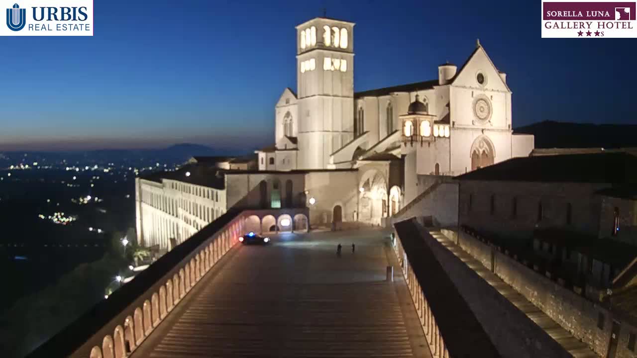 A large, illuminated basilica sits atop a hill at night, overlooking a city with lights twinkling in the distance under a dark blue sky.