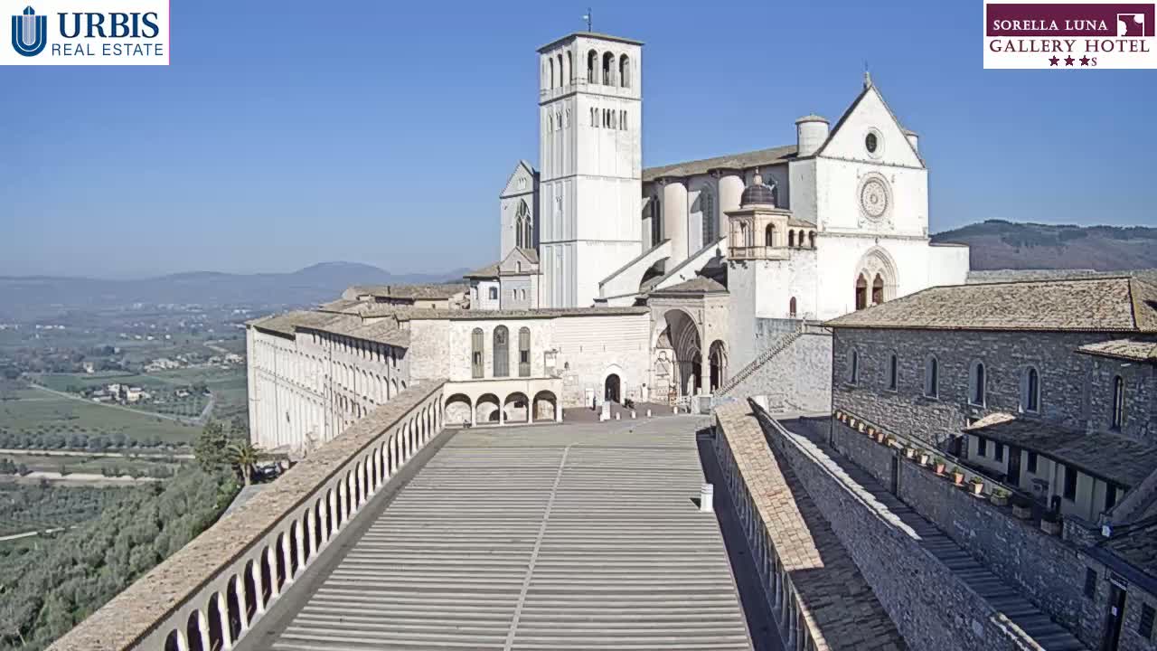 A large, light-colored stone basilica sits on a hill overlooking a valley on a clear, sunny day.