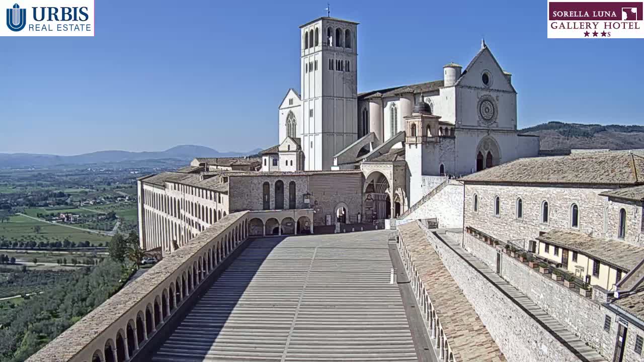 A large, light-colored stone basilica and adjoining buildings overlook a vast, paved plaza on a sunny day, with rolling hills and farmland visible in the distance.
