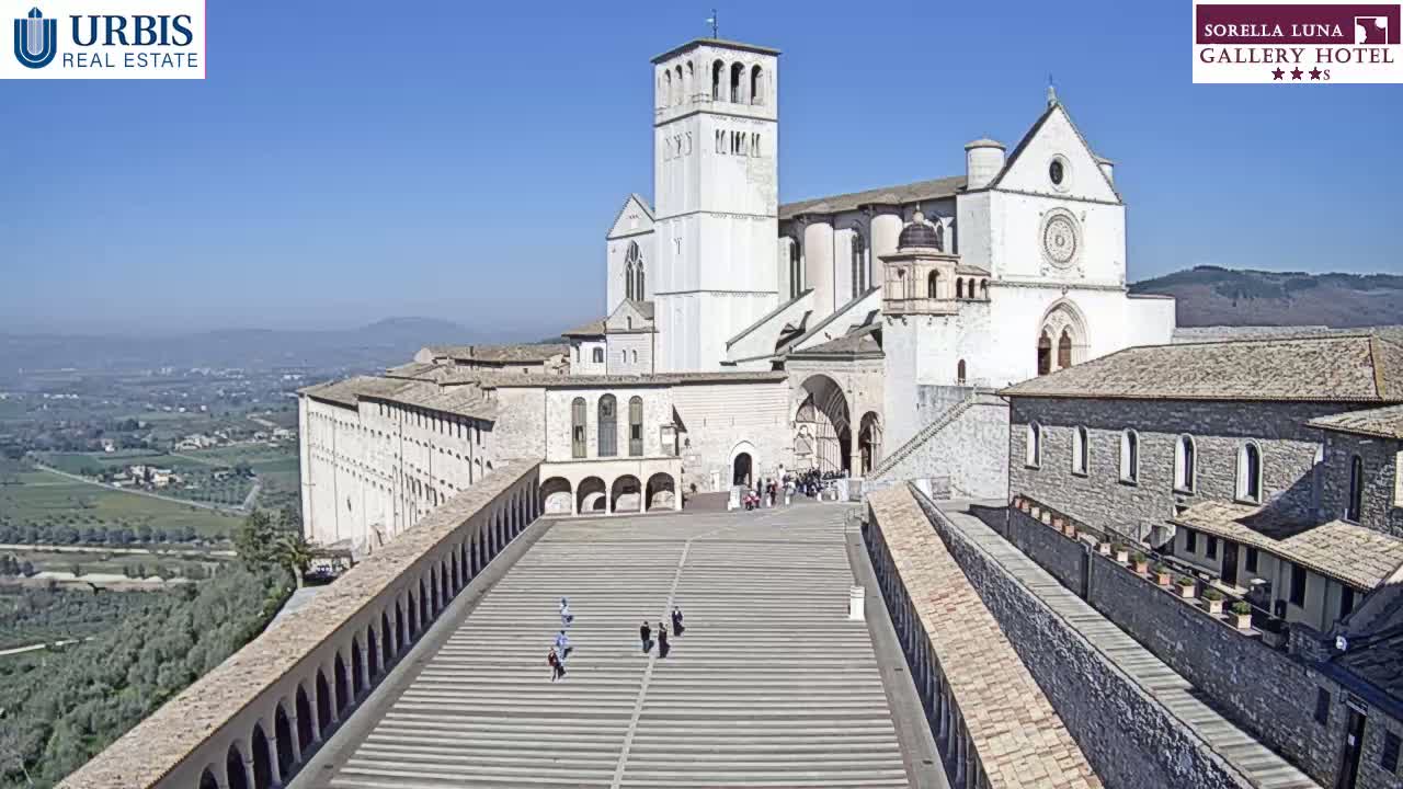 A large, light-colored basilica and adjacent buildings are situated on a hill overlooking a valley under a clear, sunny sky.