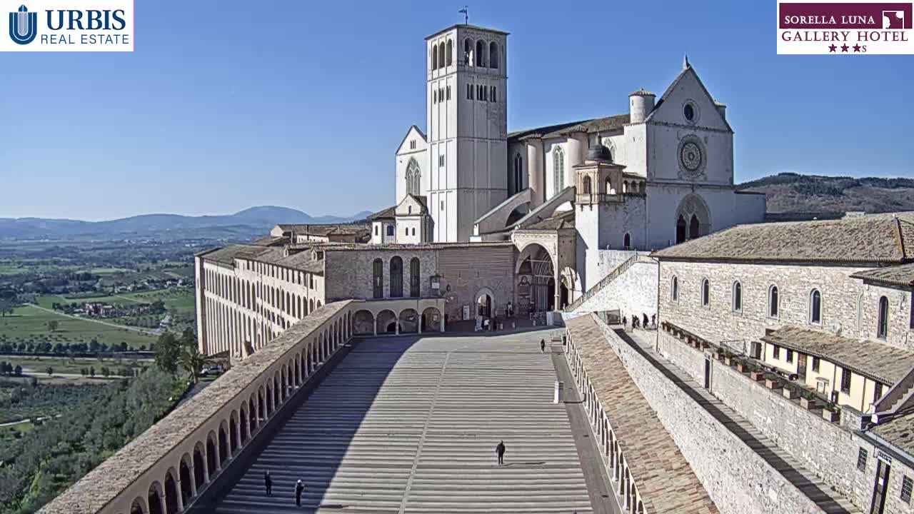 A large, light-colored stone basilica complex with a tall bell tower stands on a hill overlooking a valley on a sunny day.