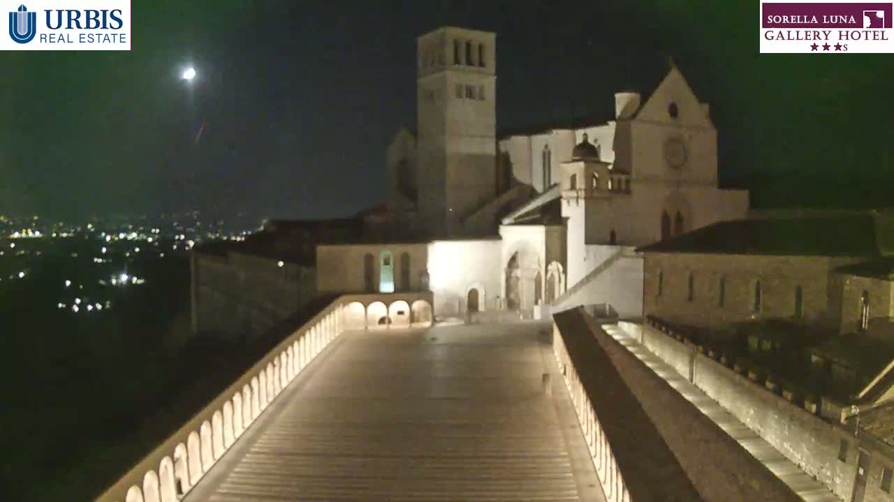 A nighttime view of a large stone building complex, illuminated, overlooking a city with a clear sky and a bright moon visible.
