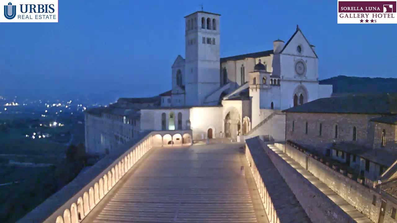 A large, illuminated basilica complex sits atop a hill at twilight, overlooking a valley dotted with distant lights.