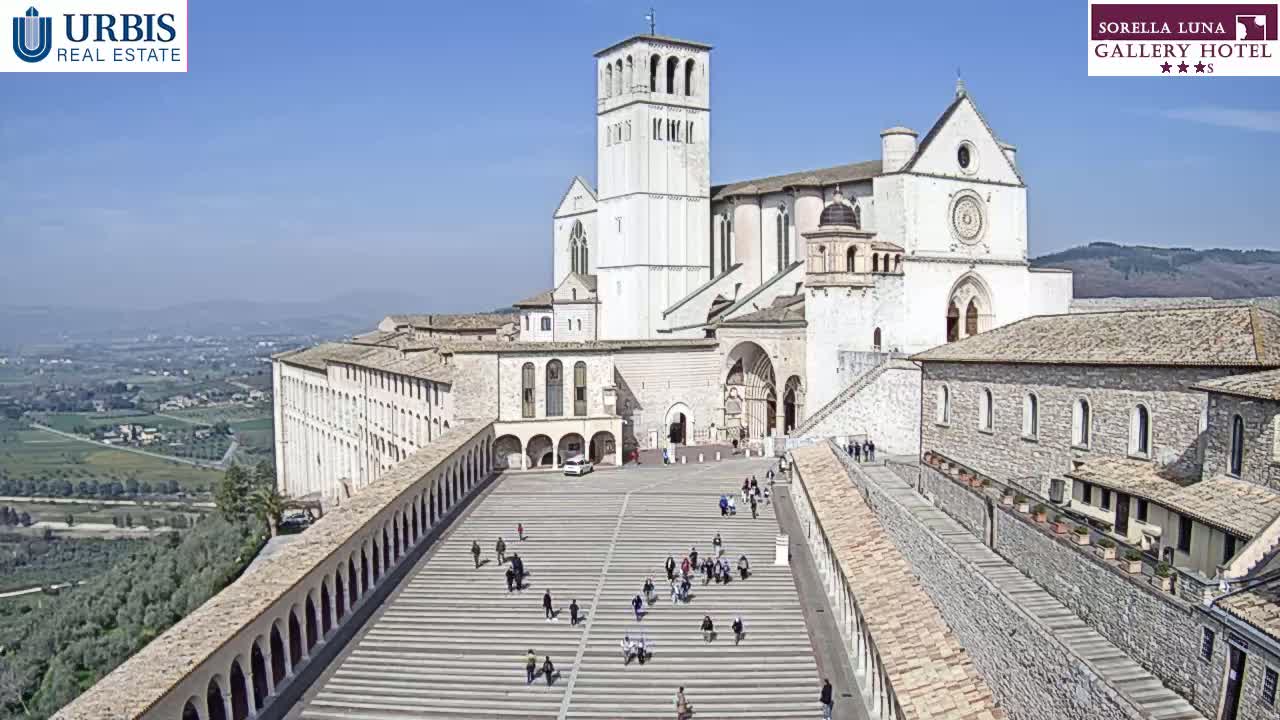 A large stone basilica and surrounding buildings are shown on a sunny day, with a paved plaza below and people walking around.