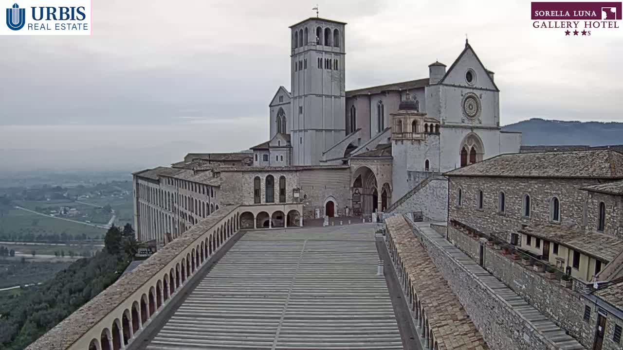 A large stone basilica and adjacent buildings are situated on a hill overlooking a valley under an overcast sky.