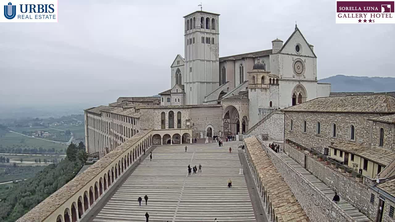 A large, light-colored stone basilica sits atop a hill overlooking a valley on an overcast day, with a wide plaza and colonnaded walkways leading up to it.