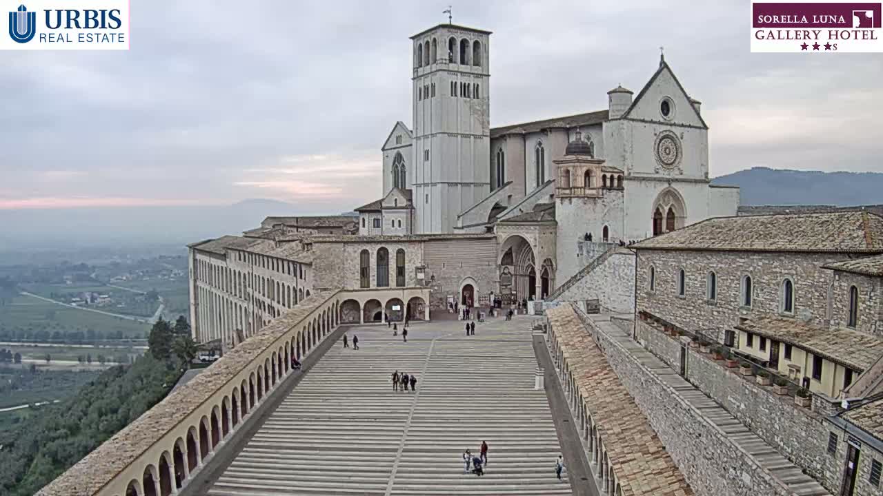 A large stone basilica and adjoining buildings are shown on a hilltop overlooking a valley on an overcast day.