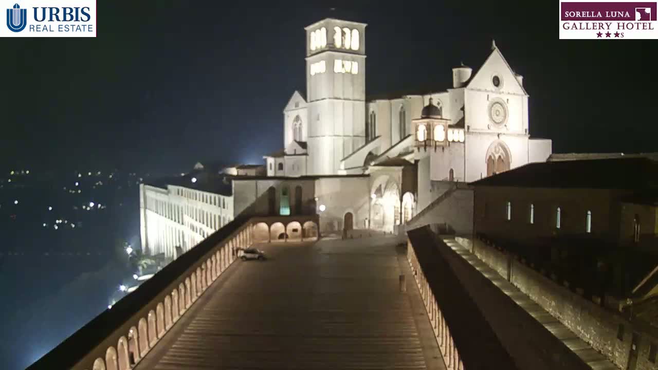 A large, illuminated basilica stands at night, overlooking a plaza and a city with distant lights.