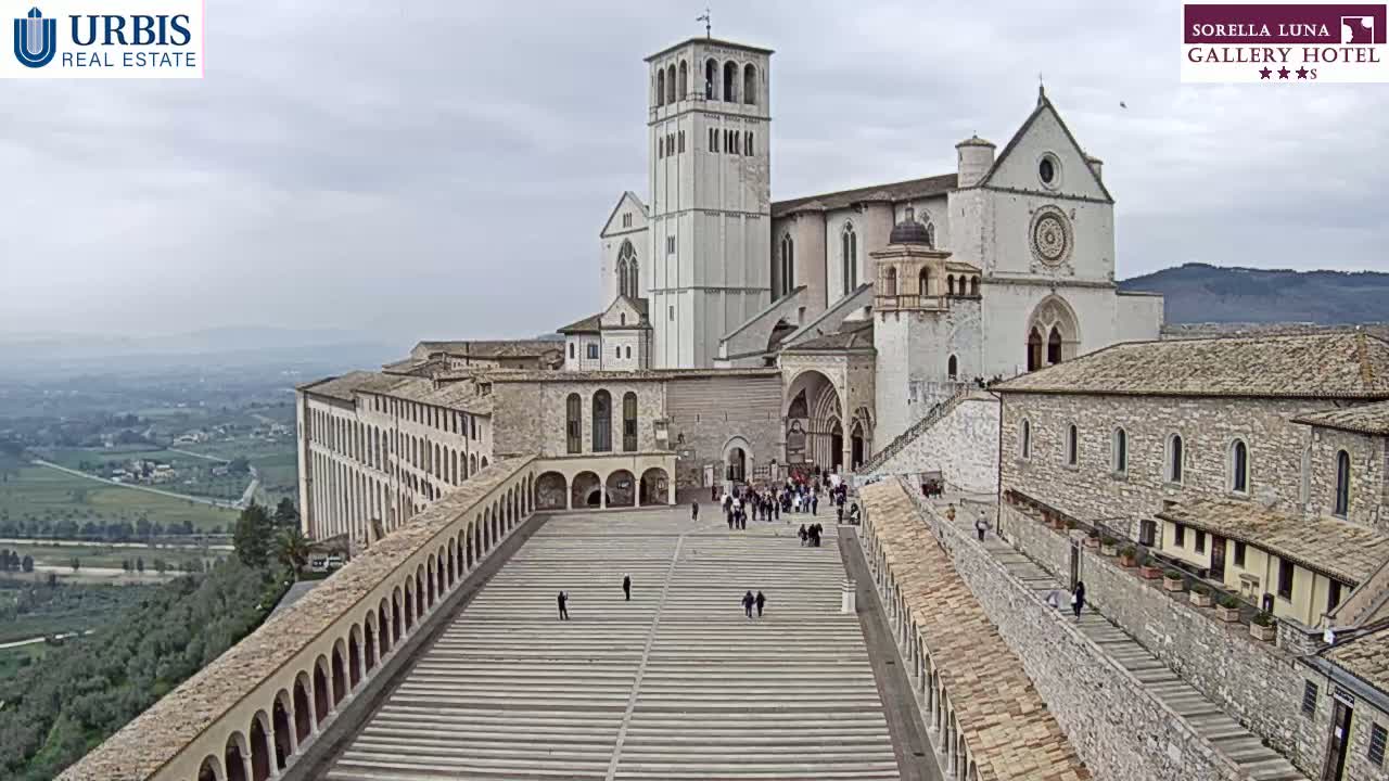 A large stone basilica and adjoining buildings are viewed from above on an overcast day, with a wide plaza stretching between them, populated by a few people.