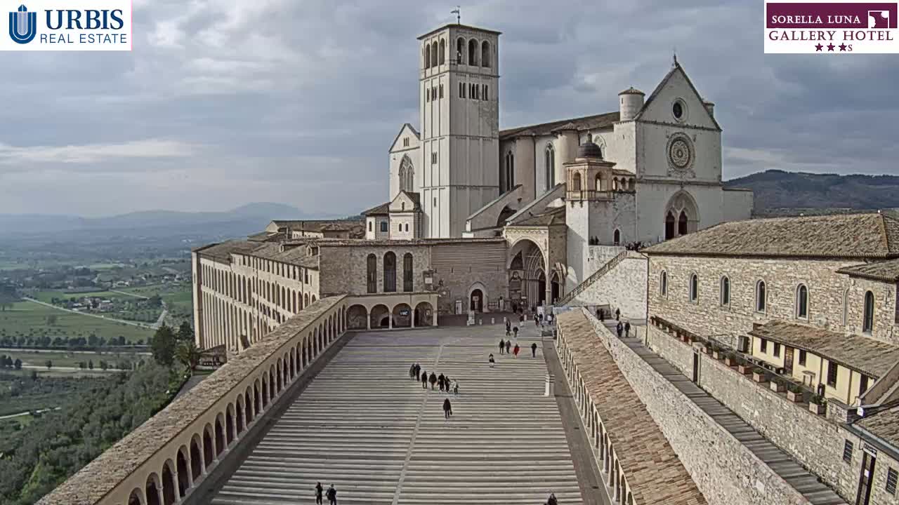 A large stone basilica and surrounding buildings are situated on a hill overlooking a valley under a cloudy sky.